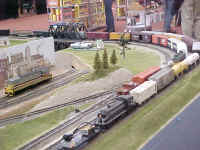 An American Models ALCO RS3 diesel locomotive in New York Cental livery pushes the train cam on a flat car past the yard entrance.